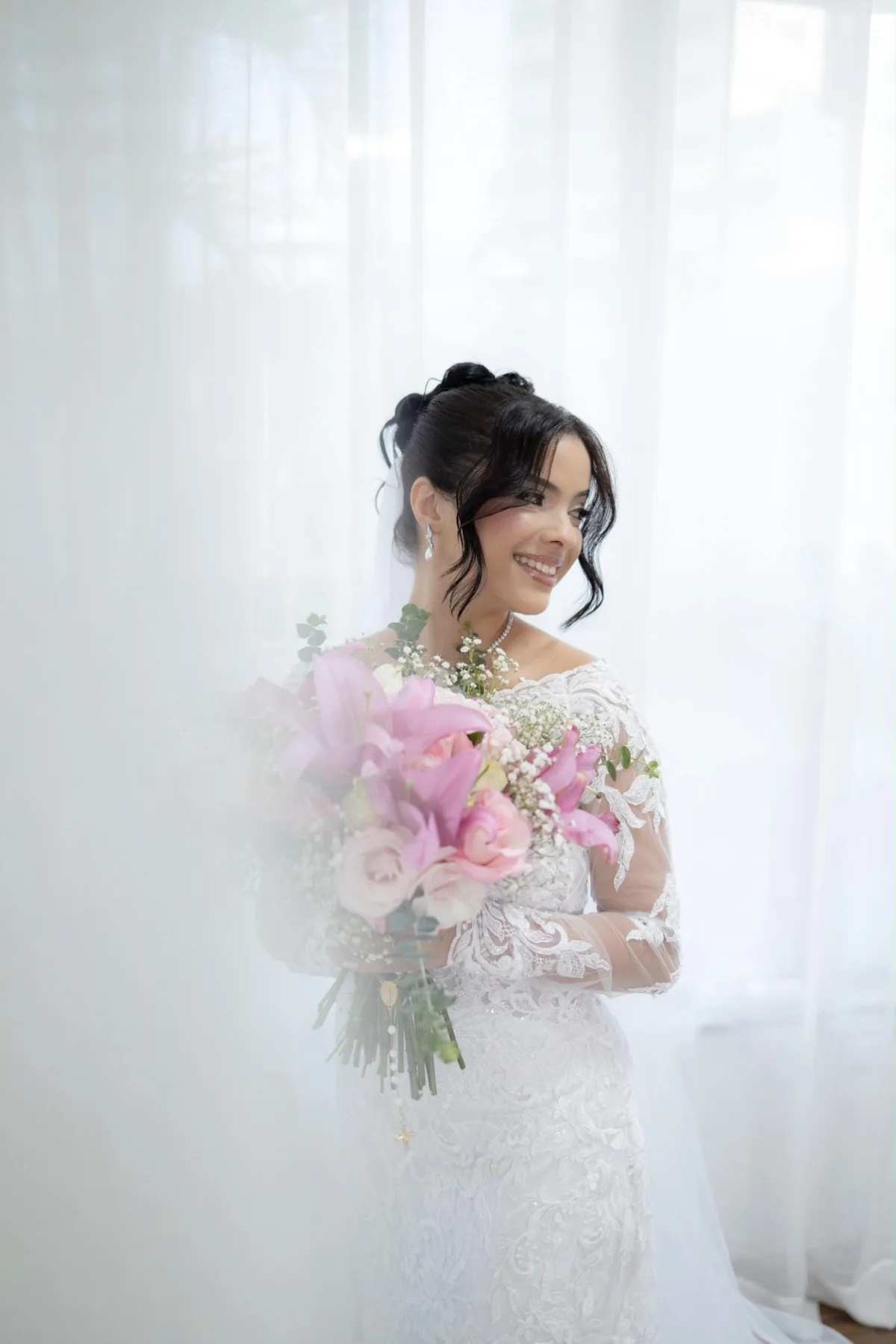 Noiva sorrindo com buquê de lírios e rosas cor-de-rosa, penteado preso e maquiagem de alta definição
